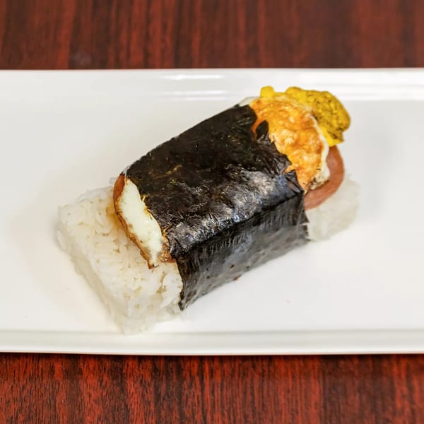 A Hawaiian Spam Musubi with rice, Spam, egg, and nori, on a white rectangular plate.